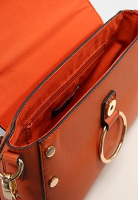 Sac à main en faux cuir orange avec une ouverture sur le dessus, des accessoires en métal doré, un accent en forme d'anneau circulaire et une poche zippée à l'intérieur, dotée d'une doublure en satin.