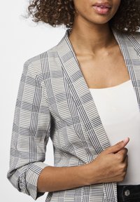 Texturovaný blazer v kostkovaném vzoru, s černobílými barvami. Design s otevřeným límcem a vyhrnutými rukávy, vrstvený přes světlejší top.