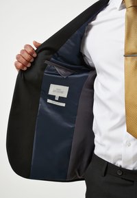 Chaqueta de traje negra, forro interior de satén azul marino, etiqueta con información de la marca y el ajuste, un bolsillo gris, textura suave, diseño ajustado.