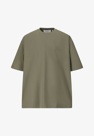 Jeune homme marchant, portant un T-shirt vert olive, un pantalon noir et des baskets blanches sur un fond blanc uni.