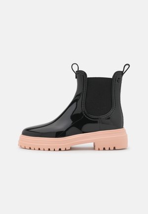 Bottes Chelsea en cuir verni noir avec une semelle en caoutchouc rose, dotées de panneaux latéraux élastiques et de languettes pour un enfilage facile. Surface texturée ajoutant de la profondeur.