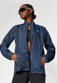 Zwarte jas met blauw geometrisch patroon, volledig met rits, hoge kraag en elastische manchetten. Draag over een zwart topje, wat een sportieve uitstraling heeft.