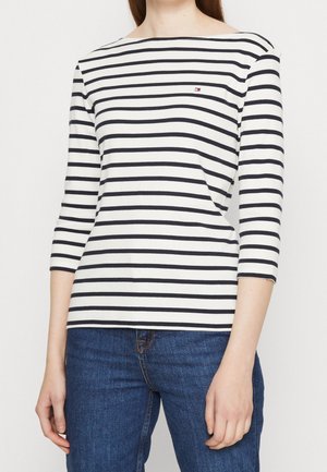 Personne portant une chemise blanche à manches longues avec des rayures horizontales noires et un jean bleu, debout devant un fond blanc uni.