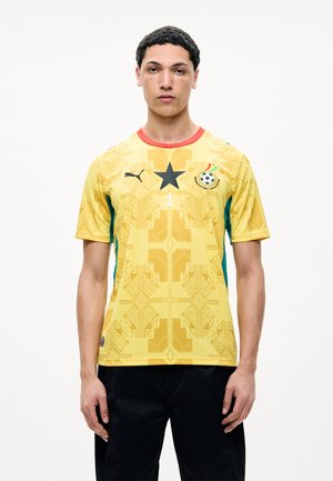 Junger Mann trägt ein gelbes Trikot der Ghana Football Association mit schwarzem Stern, Puma-Logo und Nummer 1 auf der Vorderseite, steht vor weißem Hintergrund.