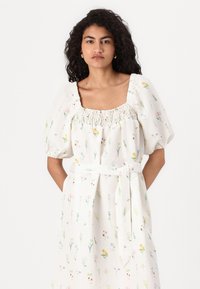 Vestido floral branco com decote quadrado, mangas bufantes, detalhes franzidos e um cinto amarrado. Padrões florais em cores pastel por toda a peça.