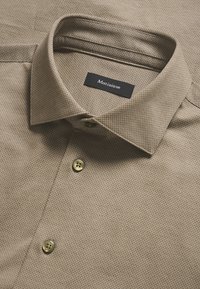 Šviesiai ruda marškinėlių chemise, pagaminta iš tekstūruotos medžiagos. Užsistatomi taškuoti apykaklė, trys auksinės mygtukai ir juoda etikete su "Martinique" užrašu.