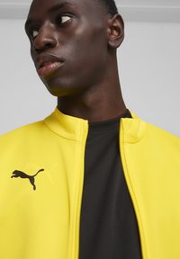 Gelbe Zip-Jacke mit hohem Kragen, ausgestattet mit einem schwarzen Puma-Logo auf der linken Brust, über einem schwarzen Shirt getragen. Glatte Textur.