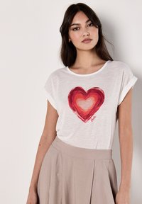 T-shirt blanc à manches courtes avec un grand graphique de cœur rouge et le texte "love", texture douce, coupe décontractée, porté avec une jupe beige.