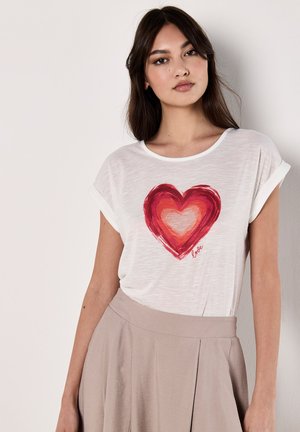 T-shirt bianco a maniche corte con una grande grafica a cuore rosso e testo "love", texture morbida, vestibilità rilassata, indossato con una gonna beige.