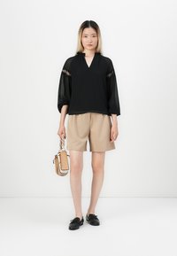 Blusa nera semi-trasparente con maniche a sbuffo, abbinata a pantaloni beige. Gli accessori includono una piccola borsa a tracolla e calzature nere.