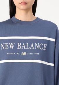 Blue sweatshirt with a ribbed crew neck. Features white horizontal stripes and “NEW BALANCE” text, along with a logo and location details.