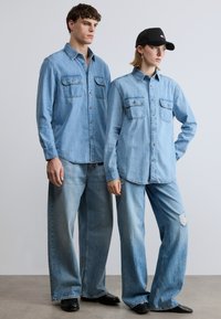 Svetlo modre jeans srajce z žepi, kombinirane z ohlapnimi svetlo modrimi kavbojkami in črno kapo. Minimalističen dizajn s sproščenim ravnim krojem.