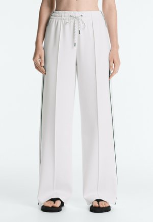 Pantaloni bianchi a gamba larga con righe laterali, vita elasticizzata con coulisse, indossati con sandali neri con punta aperta su una persona in piedi.