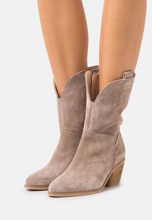 Sanden i beige mockaimid-höga westernstövlar med snedställda överkanter och staplade träklackar, bärs på bara ben.