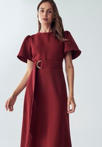 Robe brun-rouge avec des manches courtes bouffantes et une ceinture à la taille avec une boucle en or circulaire. Tissu lisse, design jusqu'au sol, silhouette simple.