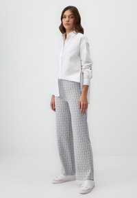 Chemise blanche à boutons, associée à un pantalon large gris clair à motifs avec de petites formes géométriques ; le mannequin porte des baskets blanches.