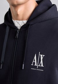 Hanorac cu fermoar de culoare bleumarin, fabricat dintr-un material moale, având un logo brodat alb "A/X" și snururi cu capete din plastic.