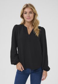 Blouse noire avec un décolleté en V, des manches longues bouffantes et des poignets élastiques. Tissu léger avec une texture lisse, associé à un jean bleu.