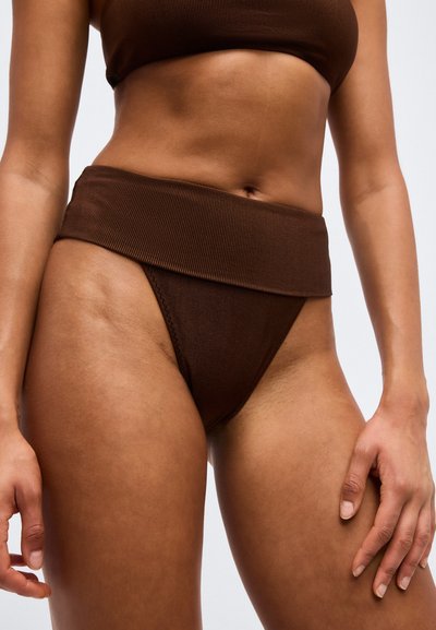 Kobieta ubrana w brązowe, prążkowane bikini z wysokim stanem i dopasowany top, odsłaniająca brzuch i górną część ud na jednolitym tle.