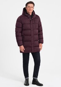 Giacca a vento bordeaux con cappuccio, caratterizzata da cuciture orizzontali, una cerniera frontale e due tasche laterali. Indossata sopra jeans scuri e stivali.