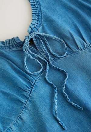 Top in denim di color blu chiaro, con colletto arricciato, chiusura a lacci e arricciature laterali. Texture morbida e liscia con leggera lucentezza.