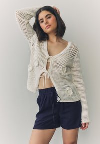 White knitted cardigan with open weave, tie closure, and textured flower accents. Paired with navy blue shorts, featuring an elastic waistband.