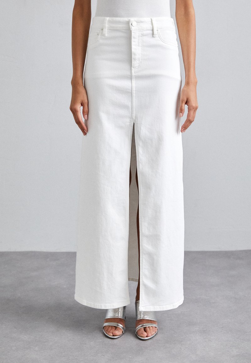 Falda maxi de mezclilla blanca con una abertura frontal, diseño de cintura alta y textura sutil. Combinada con sandalias de tiras metálicas.