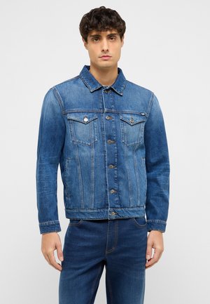 Mand iført en knappet blå denimjakke med brystlommer og matchende blå denimjeans, stående foran en ensfarvet baggrund.