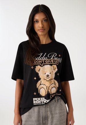 Maglietta nera oversize con maniche corte, con grafica di un orsetto pixel art, testo dorato e etichetta "Pixel Edition" in basso.