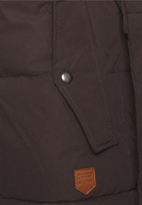 Brown insulated jacket with a quilted texture, featuring a metal snap button and a tan leather logo patch on the lower right corner.