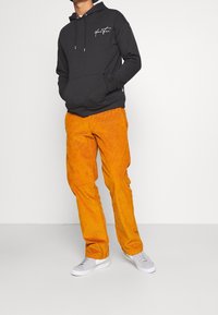 Sudadera negra con un bolsillo frontal y un logo blanco, combinada con pantalones de pana naranjas y zapatillas grises. Materiales texturizados y contraste de colores atrevidos.