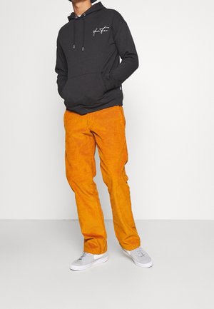 Felpa nera con tasca frontale e logo bianco, abbinata a pantaloni in velluto a coste arancioni e sneakers grigie. Materiali texturizzati e contrasto di colori audaci.