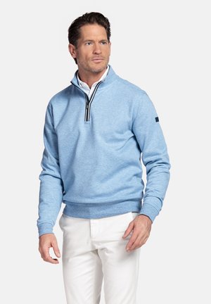 Man draagt een lichtblauwe trui met een korte rits over een wit overhemd en een witte broek, staat tegen een egale lichte achtergrond, kijkt naar rechts.