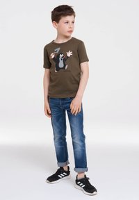 Camiseta verde oliva con un personaje de topo de dibujos animados sosteniendo una pequeña pala, combinada con unos vaqueros de denim azul con puños enrollados y zapatillas negras.
