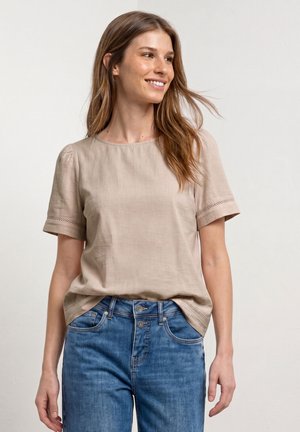 Femme souriante portant un chemisier beige à manches courtes avec bordure en dentelle et un jean bleu taille haute sur fond uni.
