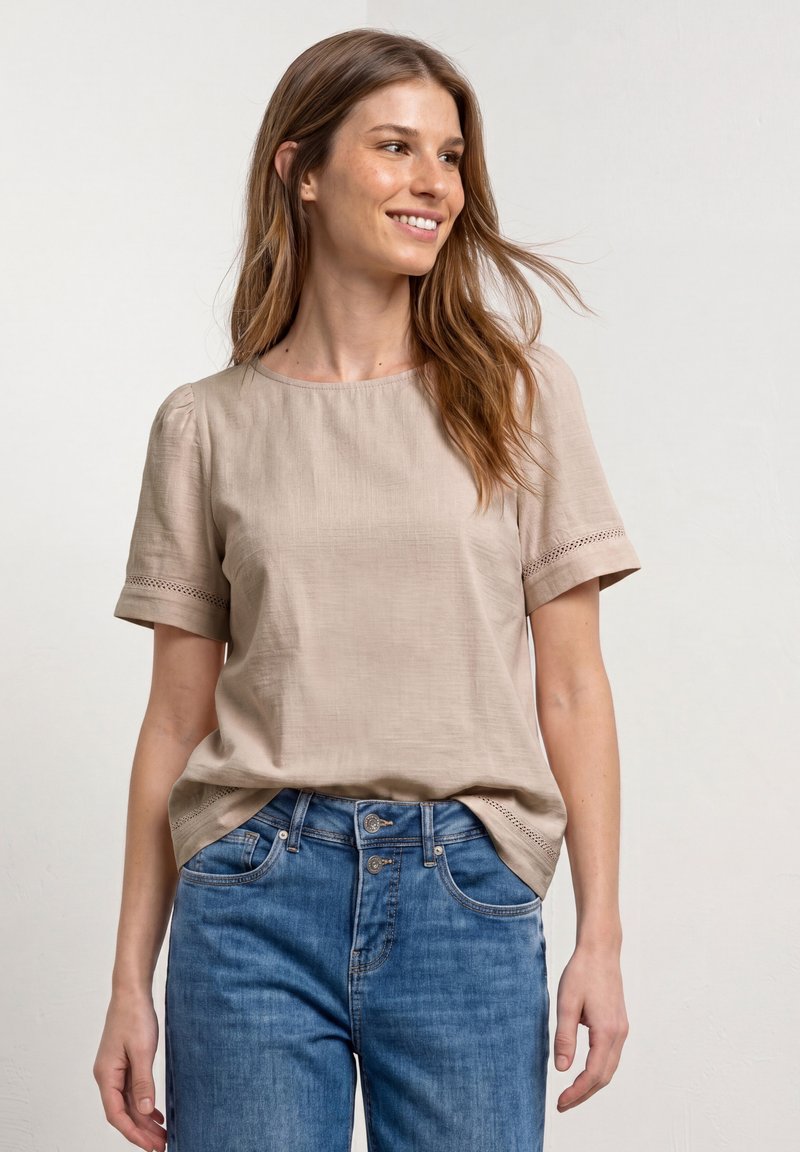 Femme souriante portant un chemisier beige à manches courtes avec bordure en dentelle et un jean bleu taille haute sur fond uni.