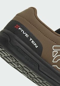 Ruskea urheilukenkiä mustilla yksityiskohdilla; siinä on teksturoitu pinta, kumipohja, jossa on ulkonevia pitoja, sekä "Five Ten" -brändäys valkoisena ja punaisena.