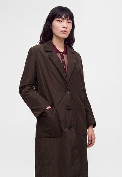 Femme aux longs cheveux foncés porte un manteau marron foncé boutonné par-dessus une chemise bordeaux à col, debout devant un fond clair uni.