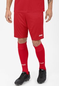 Rote Sportshorts und passende kniefreie Socken, beide aus glattem, synthetischem Material. Schwarze Fußballschuhe mit strukturiertem Design.