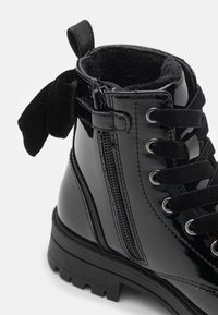 Bottines en cuir verni noir avec une doublure en fausse fourrure, un laçage à l'avant, une fermeture éclair sur le côté et un détail de nœud en tissu au col. Semelle texturée.