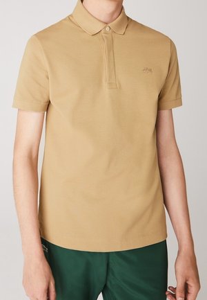 Beige polo met korte mouwen, kraag en knopen, gedragen door een persoon met groene korte broek, met het Lacoste krokodillenlogo op de borst.
