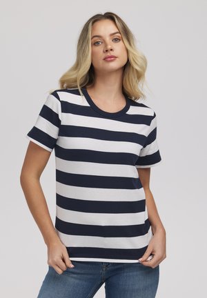 Kobieta o blond włosach, ubrana w granatowo-biały t-shirt z poziomymi paskami i krótkim rękawem oraz niebieskie jeansy, stojąca na szarym tle.