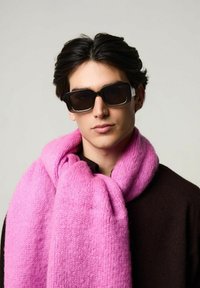 Lunettes de soleil noires, écharpe rose avec une texture douce, portée sur un pull marron foncé. L'écharpe a un motif en maille épaisse.