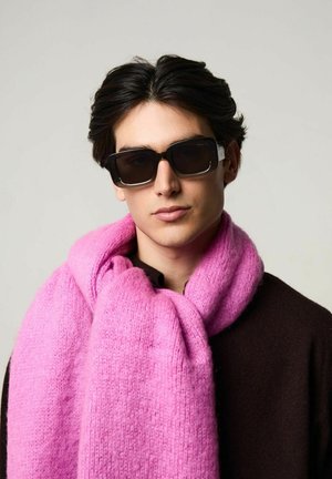 Lunettes de soleil noires, écharpe rose avec une texture douce, portée sur un pull marron foncé. L'écharpe a un motif en maille épaisse.
