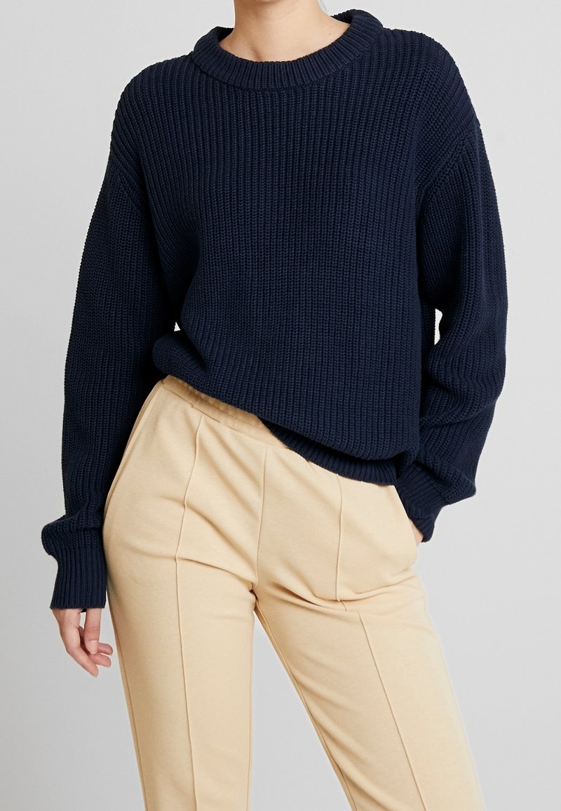 Maglione lavorato a maglia blu navy con texture a coste, dotato di un ampio scollo rotondo e una vestibilità rilassata, abbinato a pantaloni beige a gamba affusolata.