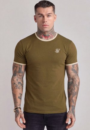 Mand med tatoveringer på nakke og arme iført olivengrøn tætsiddende t-shirt med beige kant, stående mod en ensfarvet lys baggrund.
