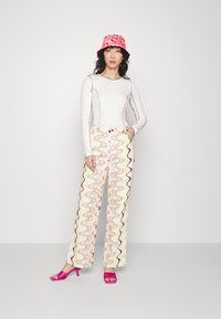 Top branco de mangas compridas com costuras contrastantes, combinado com calças de perna larga apresentando padrões ondulados multicoloridos. Chapéu de balde rosa e saltos altos.