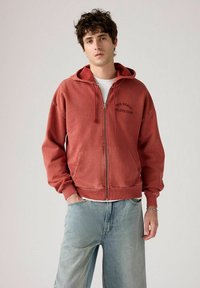 Felpa rossa con zip, dotata di tasca frontale e cappuccio con stringa. Presenta un logo stampato sopra la tasca. Indossata con jeans blu a vestibilità ampia.