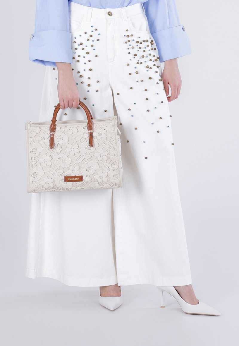 Vrouw draagt witte broek met wijd uitlopende pijpen en studs, lichtblauw overhemd, witte hakken, en houdt een beige kanten handtas met bruine handvatten vast.