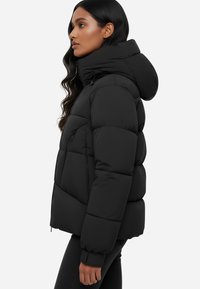 Giacca a vento nera imbottita con design trapuntato e colletto alto. Presenta una tasca con zip sul lato e polsini aderenti. Texture liscia e opaca.